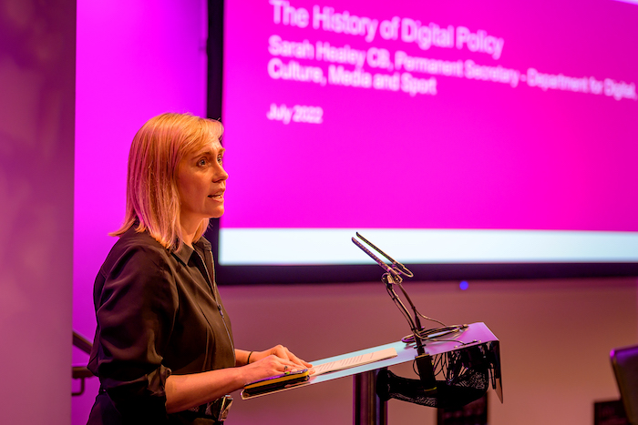 Sarah Healey standing at a lectern, in profile, with an indistinct powerpoint presentation on display in the background